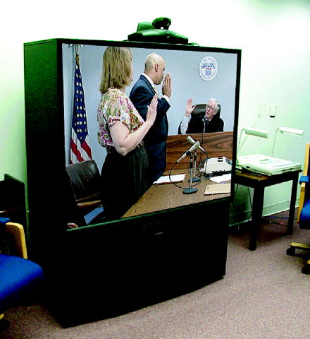 This photo is of a large screen television.  The screen shows a man and woman standing and raising their right hands and a judge sitting and raising his right hand.  