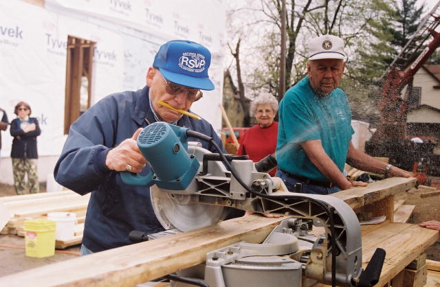 Senior volunteers are using a power saw to cut boards for a Habitat for Humanity project.  