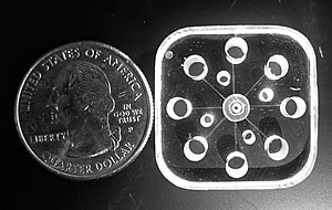 Photo showing the core of the new NIST miniature GEMBE chemical separation device, a machined acrylic block, with a quarter for scale. Eight sample reservoirs for multiplexed separations form a ring around the central buffer solution port.