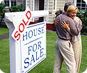 A man and woman hugging in front of their new home; the 'house for sale' sign has 'sold' over it.