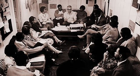 A man sits at one end of a narrow rectangular room and speaks to the assembled group that sits on chairs arranged against the walls.