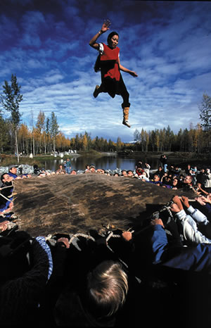 A young woman is suspended in the air against a backdrop of an open sky filled with clouds.  Below is the tense surface of a raw hide tranpoulin pulled by  many hands.