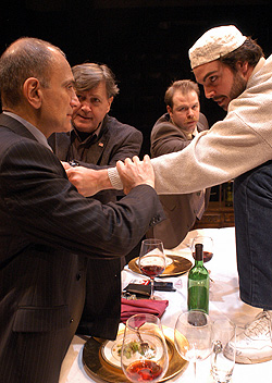 A young man dressed in white faces three older men in suits across a dinner table.  The table is strewn with the remains of a meal-- half-empty wine glasses, an empty wine bottle, used dinner plates.  In the forefront of the scene, the younger man grabs the suit lapel of one of the older men, while the older man grabs the younger man's arm. In the background,  the two remaining men try to push the older man and younger man apart.