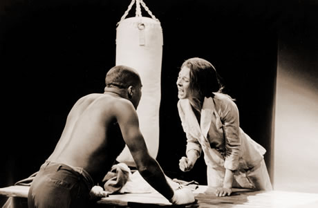 Photograph of an African-American man and a White woman facing each other in a heated exchange.  A table is situated between them and a boxing punching bag can be seen in the background.