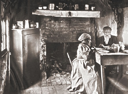 Two older people, a man and a woman are seated at a table having supper.  At the center there is a fireplace, at the left a small cupboard ans a window.  The man is dressed in black and has a long white beard, the woman is has a long dress and wears a headscarf