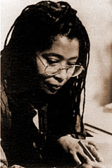 This is a headshot of African American author Alice Walker. In the photo, Walker wears glasses and her hair is in long braids. She's looking down, perhaps at a sheet of paper, while holding a pen in her left hand.