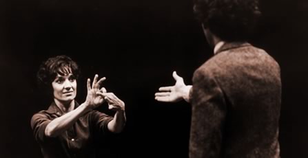 Two actors on stage.  A man seen from the back, a woman facing him signals with her hands in sign language