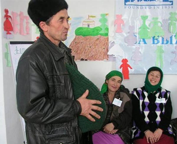 Two women in background smile as man stands at front of classroom, hands resting on sweater stretched out by sympathy belly that makes him appear pregnant 