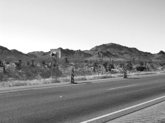 The cover photograph shows the monitoring equipment used to calculate travel times through the work zone. It shows a small cross-section of the two-lane road along with two small pole-mounted signs on the other side of the road, each protected by a striped cone. There is a camera mounted on the back of the first sign, and a light mounted on the back of the second sign. Mountains can be seen in the distance.