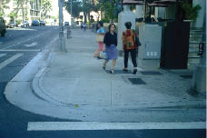 Case Study: Two contrasting photos show a US intersection with space-consuming flared curb ramps compared with an intersection in Barcelona, Spain, where the sides of the ramps are shielded from pedestrian travel by poles or trash cans, leaving more sidewalk area for pedestrians.