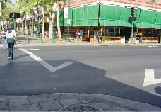 Case Study:  Two photo views of the same urban intersection show how an existing storm sewer inlet precludes the addition of a curb ramp within the crosswalk on one side of the crossing