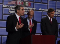 Secretary Gutierrez speaking at podium with Secretary Chertoff and Canadian Minister of Industry Maxime Bernier at his side.