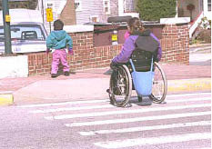 Pedestrian using a wheelchair approaching a curb ramp from a public street crosswalk