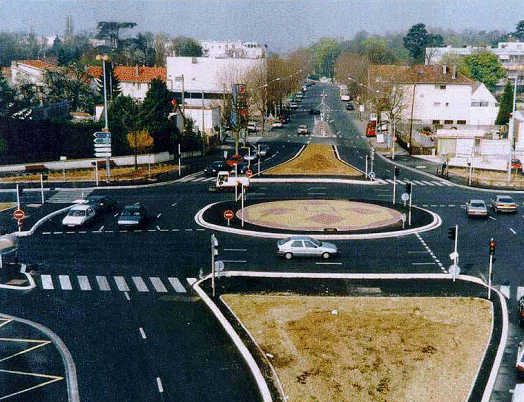 Figure 5.  Photo.  Signalized roundabout in France where crosswalks are located adjacent to the circular roadway.  Note that France only allows post mounted signals.  It is possible that overhead signals may be used in the US.  This photograph depicts a large triple-lane roundabout in France.  Post mounted signals are located on both side of the roadway adjacent to clearly marked stop bars.  Operationally, this roundabout functions more like a standard intersection because of the signals.  However, it does incorporate geometric design features found in roundabouts, namely a center island and deflection on the entry and exit.