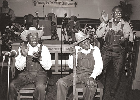 Three Senior African Americans clap and sing, two of them are seated and one is standing at a microphone. A sing hangs in the background with the inscrition: Welcome, New Zion Missionary Baptist Church.