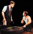 Man standing talking to another man squatting working on a large truck tire