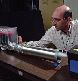 Photo of researcher in lab using small, square box that blows smoke through a long, metallic tube into a hybrid vehicle battery pack to study air flow within the pack.