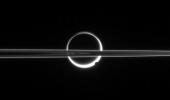 Saturn's rings cut across an eerie scene that is ruled by Titan's luminous crescent and globe-encircling haze, broken by the small moon Enceladus, whose icy jets are dimly visible at its south pole. North is up