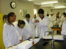 The photograph shows four minority students conducting research in a laboratory at the University of North Carolina at Pembroke.  The student on the right is using a pipette while the others three students are observing. Three of the students wear safety goggles. All are wearing white lab coats.
