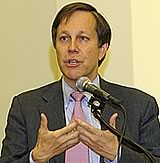 Photo of Dana Gioia, Chairman of the National Endowment for the Arts. He wears a gray suit, light blue shirt with pink tie and speaks into a microphone. He gestures with both his hands elevated, upturned and intertwined.