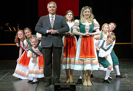     Middle aged man on stage in a suit and tie with eight young boys and girls dressed in period outfits  						