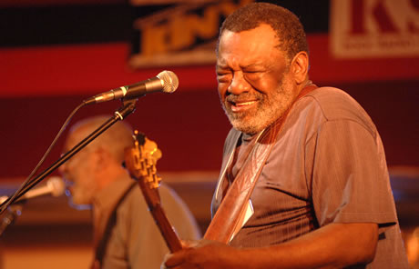 A musician in front of a microphone, his eyes closed tightly, in a gesture of concentration.  A guitar in his hands is partially visible.		