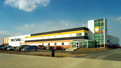 Photo of a warehouse-like large rectangular two-story building, with the words 'First Solar' on its side and on a taller stairwell mounted on one side of the building. A glass entrance atrium is at one corner and a number of vehicles are parked in a lot in front of the building.