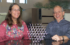 Photo of a smiling man and woman, both wearing glasses, sitting at a table upon which sits a model of a crystal array. The woman has long hair and the man has short graying hair.