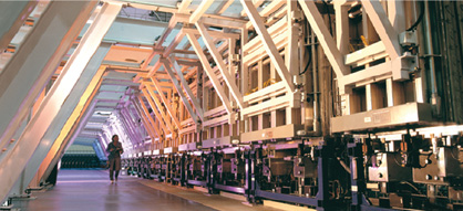 Photo of a long line of heavy industrial equipment along an A-shaped corridor framed by steel girders. A woman is walking down the corridor, which recedes into the distance.