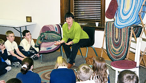 A woman sitting on a chair talks to children sitting in a circle on the floor.  In the background examples of her rug work can be seen.		