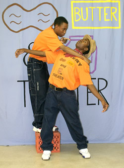 Two young actors on stage, one standing on a small box holds the other by his shoulders forcing him to lean back, the backdrop has words written on it: Peanut, butter and has the drawing of a jar		