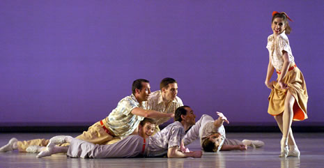 A smiling female dancer, wearing a poodle skirt, poses flirtatiously while a group of five male dancers, in khakis and flowered shirts, lie on stage reaching toward her		
