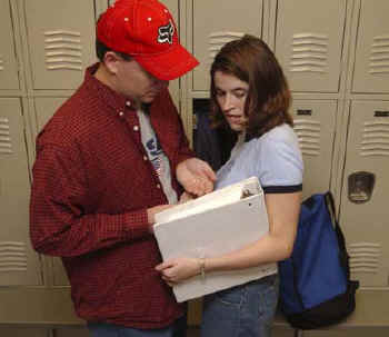 Picture of a male and female in front of lockers using a large binder to shield a drug purchase. 