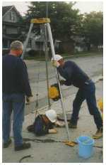 A person being lowered into a manhole to collect water samples for monitoring.