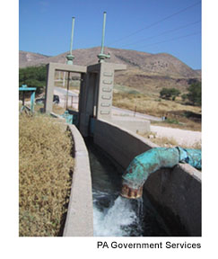 A pipe delivers water into a cement irrigation canal in a dry landscape with hills in the background. Photo Source: PA Government Services