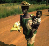 Photo of a woman holding a young child, while balancing items on her head, walking down the road.