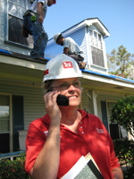 Photo of a lady talking on a phone in front of a house with construction workers on the roof.