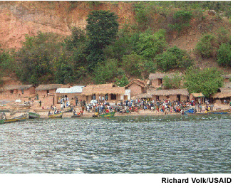 A small fishing village can be seen at the base of low cliffs. People have gathered on the seashore among fishing boats that have been beached there. Photo Source: Richard Volk/USAID