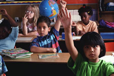 Image: A group of young school children raising their hands in class.