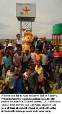 Pictured from left to right, back row:  Robert Knezevic, Project Director for Takalani Sesame; Kami, the HIV-positive Muppet from Takalani Sesame; U.S. Ambassador Eric M. Bost; Trevor Field, PlayPumps inventor;  and local children at a school ground  in South Africa that depend on the merry-go-round style water pump.