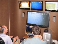 Inside the container, pilot, Matthew Cook (left), directs the ROV on the seafloor while he watches real-time video on the large screen monitor. Geoff Cook (right), co-pilot, prepares to maneuver the manipulator arm for sample collections. The top screens include information on the ROV position and direction, digital still camera views, and position of the ROV relative to the ship (far right screen). - (photo credit: USGS DISCOVRE Expedition) - click to enlarge