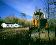 [photo:] Cleared forest lot with new single story white residential home in background and bulldozer in foreground.