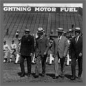 Four men in suits walking across a baseball field, followed by baseball players.