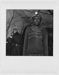 Unidentified miner, half-length portrait, facing front, wering headlamp, in mine with two miners in the background, Zimbabwe