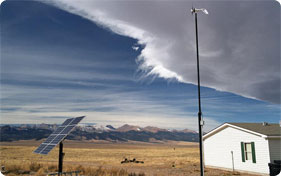 Solar panels and a small wind turbine are to the left of a one-story house on the prairie, with mountains in the background.