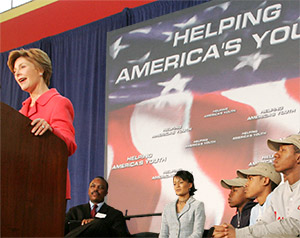 Photo of Mrs. Laura Bush standing behind a podium on a stage with a Helping America's Youth banner on the wall behind her. There are several young adults seated behind her to her left.