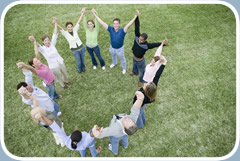 A group of twelve men and women stand outside in a circle. They are holding hands with their arms stretched into the air.
