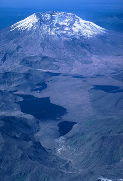 Color photo: Aerial view of snow-capped new mountain with Spriit Lake and a baren landscape in the foreground.