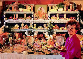 Photo: A smiling woman stands before a many tiered display of food , candles, family portraits and religious artifacts.