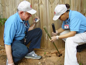 Photo: two persons kneeling near a fence.  One has a stick that is in a hole in a ground.